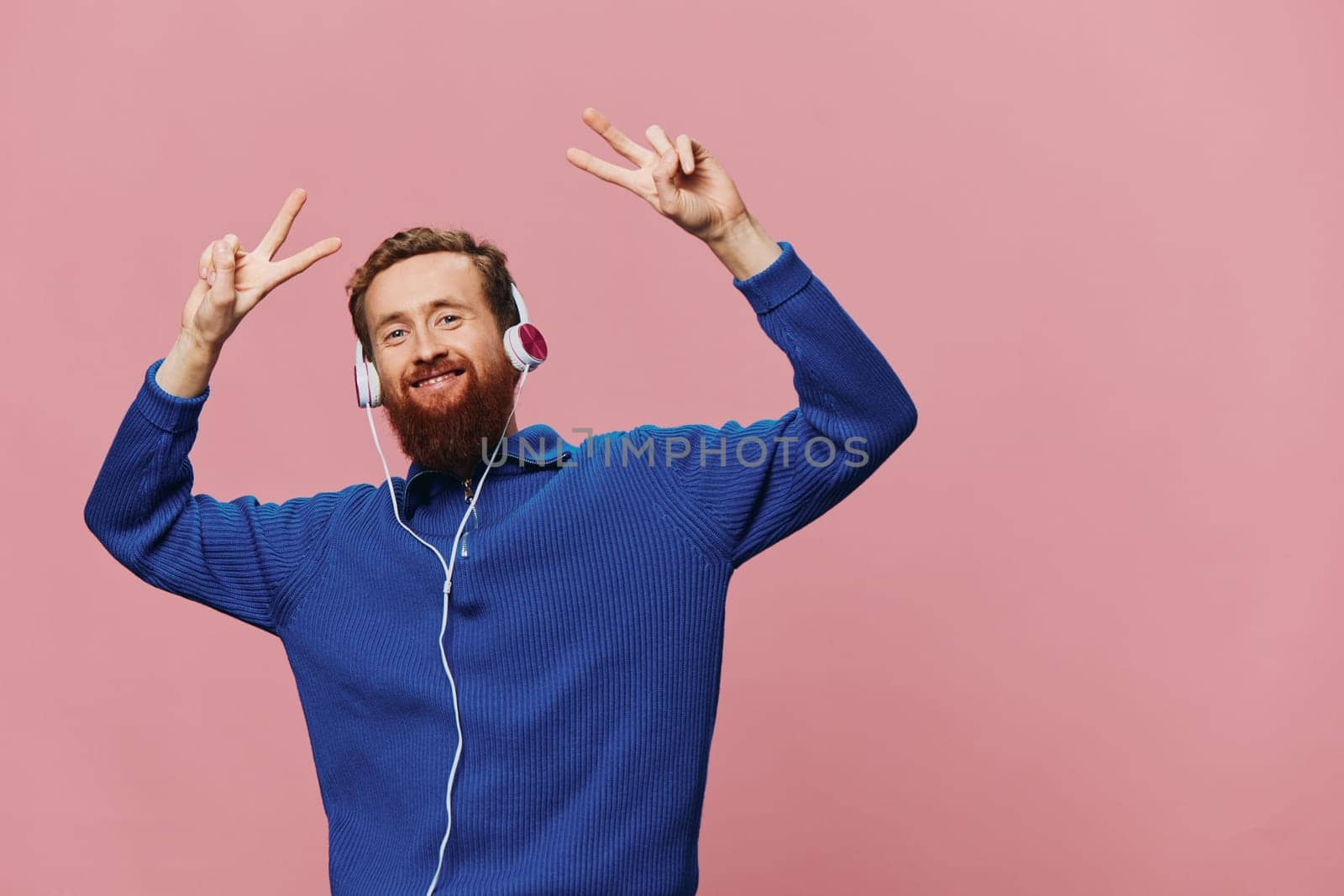 Portrait of a redheaded man wearing headphones smiling and dancing, listening to music on a pink background. A hipster with a beard. High quality photo