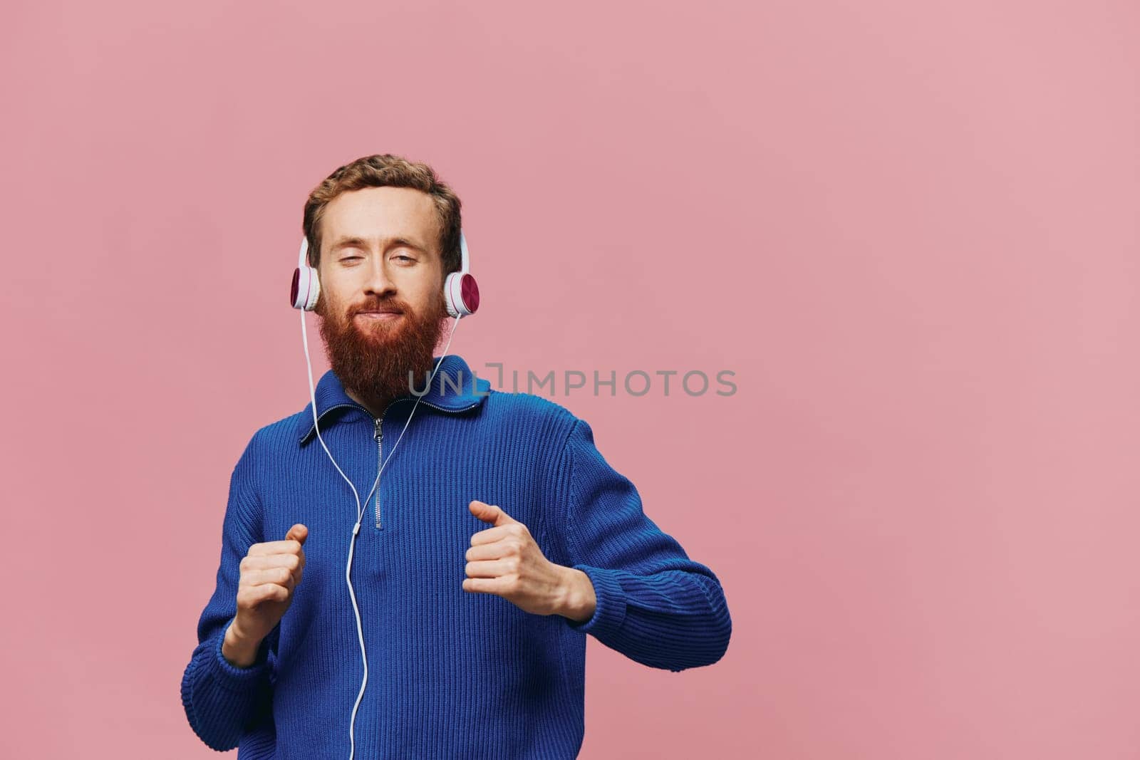 Portrait of a redheaded man wearing headphones smiling and dancing, listening to music on a pink background. A hipster with a beard. High quality photo