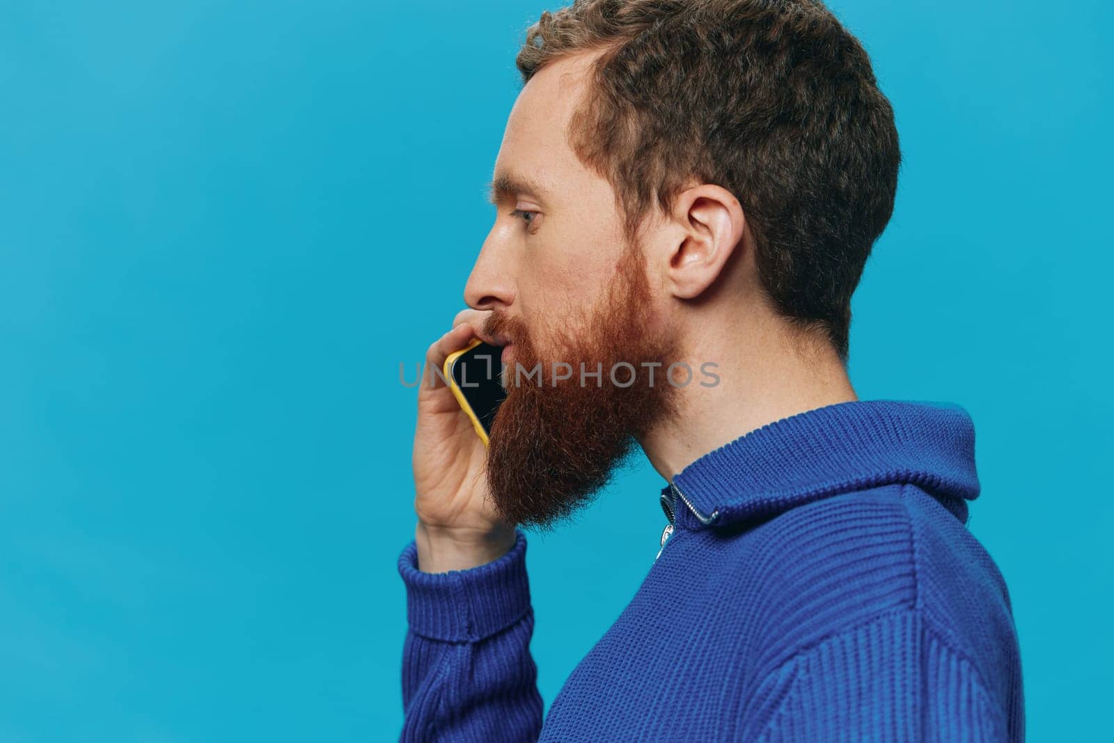 Portrait of a man with a phone in his hands does looking at it and talking on the phone, on a blue background. Communicating online social media, lifestyle by SHOTPRIME