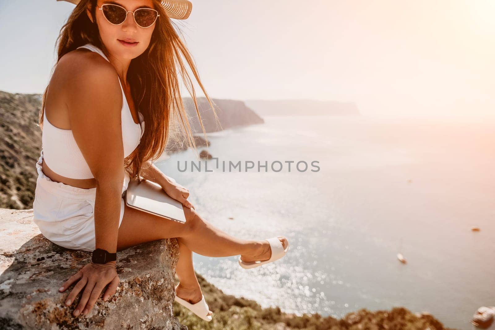 Successful business woman in yellow hat working on laptop by the sea. Pretty lady typing on computer at summer day outdoors. Freelance, travel and holidays concept.