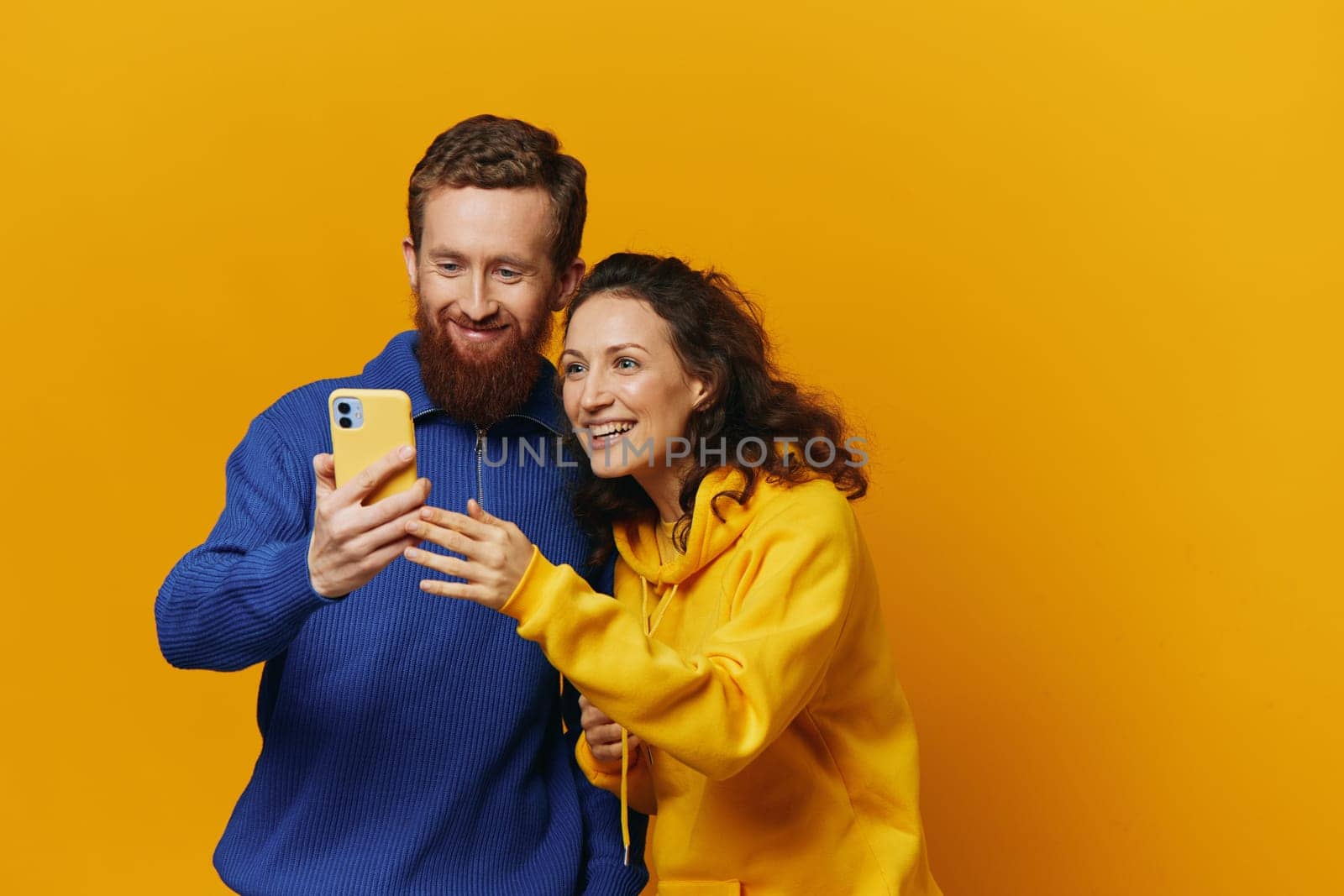 Man and woman couple smiling merrily with phone in hand social media viewing photos and videos, on yellow background, symbols signs and hand gestures, family freelancers. by SHOTPRIME