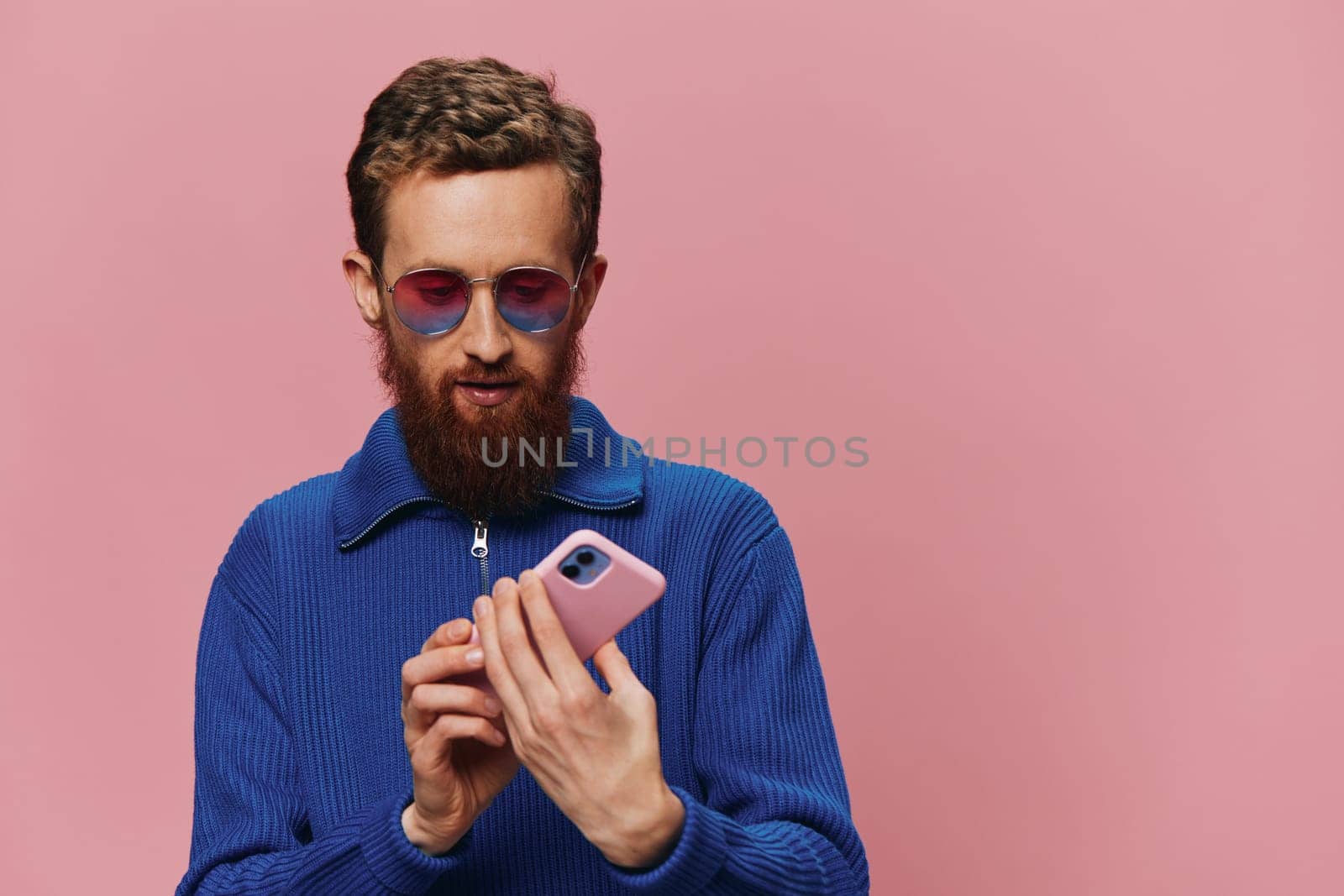 Portrait of a redheaded man with phone in hand taking selfies and photos on his phone with a smile on a pink background, blogger. High quality photo