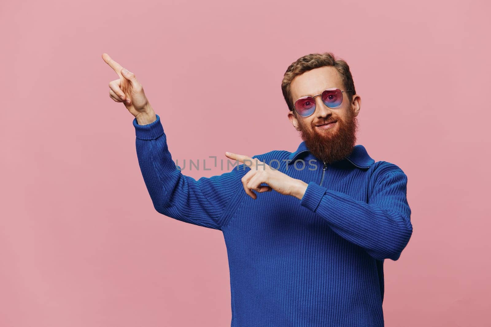 Portrait of a redheaded man wearing sunglasses smiling and dancing, listening to music on a pink background. Hipster with a beard, happiness finger pointing. High quality photo