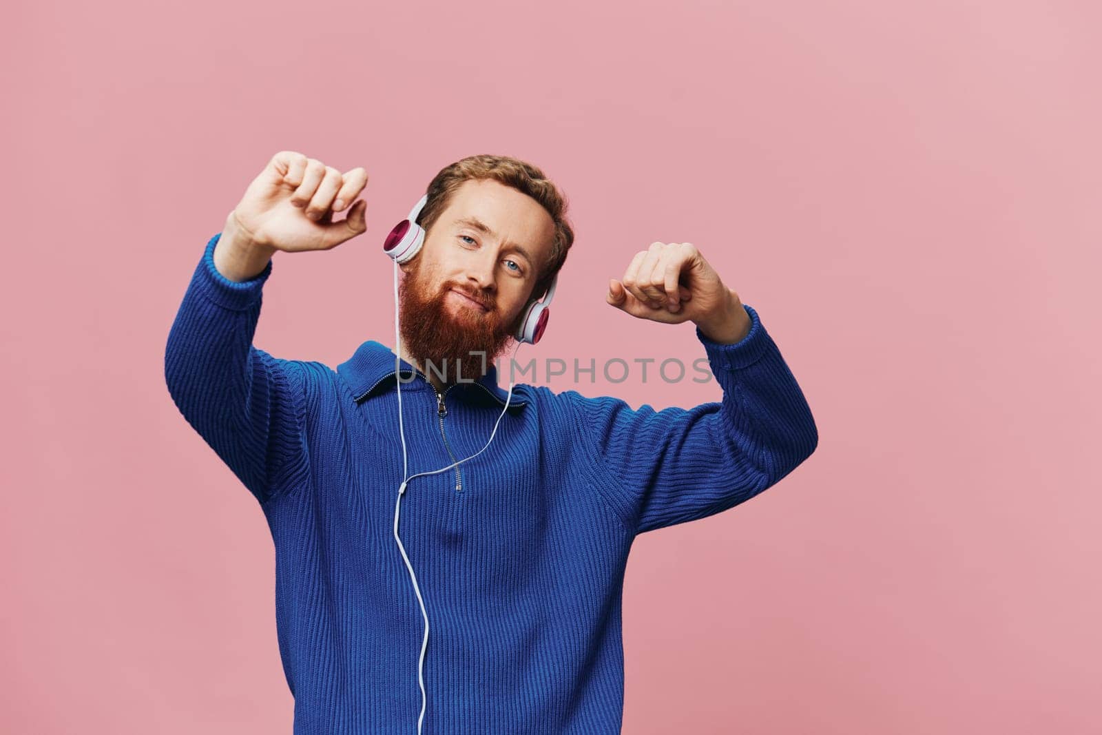 Portrait of a redheaded man wearing headphones smiling and dancing, listening to music on a pink background. A hipster with a beard. High quality photo