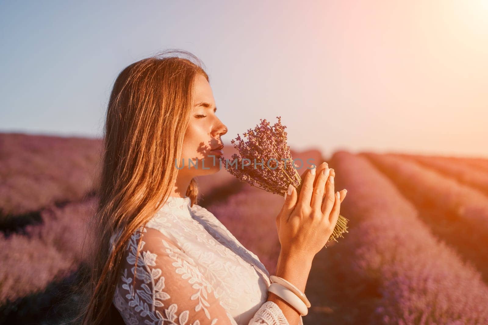 Close up portrait of young beautiful woman in a white dress and a hat is walking in the lavender field and smelling lavender bouquet.