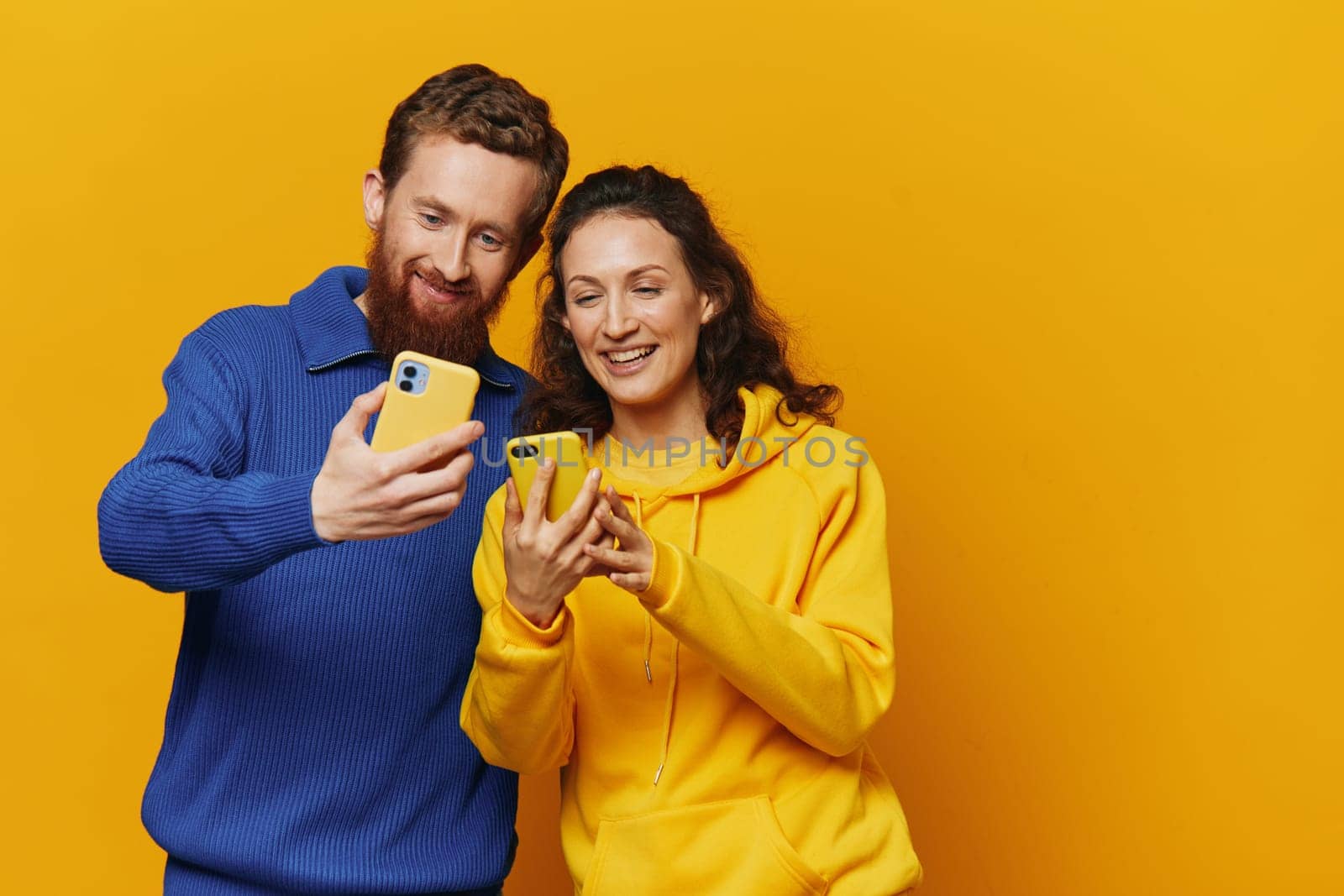 Man and woman couple smiling merrily with phone in hand social media viewing photos and videos, on yellow background, symbols signs and hand gestures, family freelancers. by SHOTPRIME