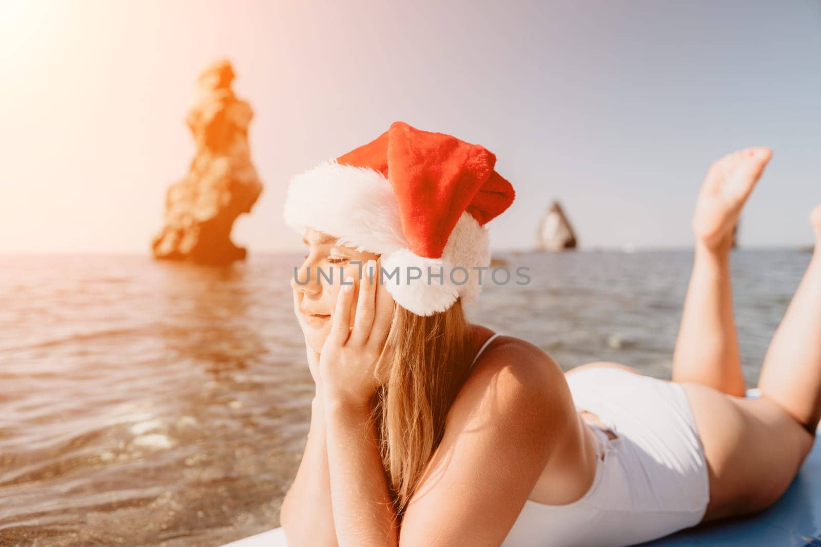 Woman sea sup. Close up portrait of happy young caucasian woman with long hair in Santa hat looking at camera and smiling. Cute woman portrait in a white bikini posing on sup board in the sea by panophotograph