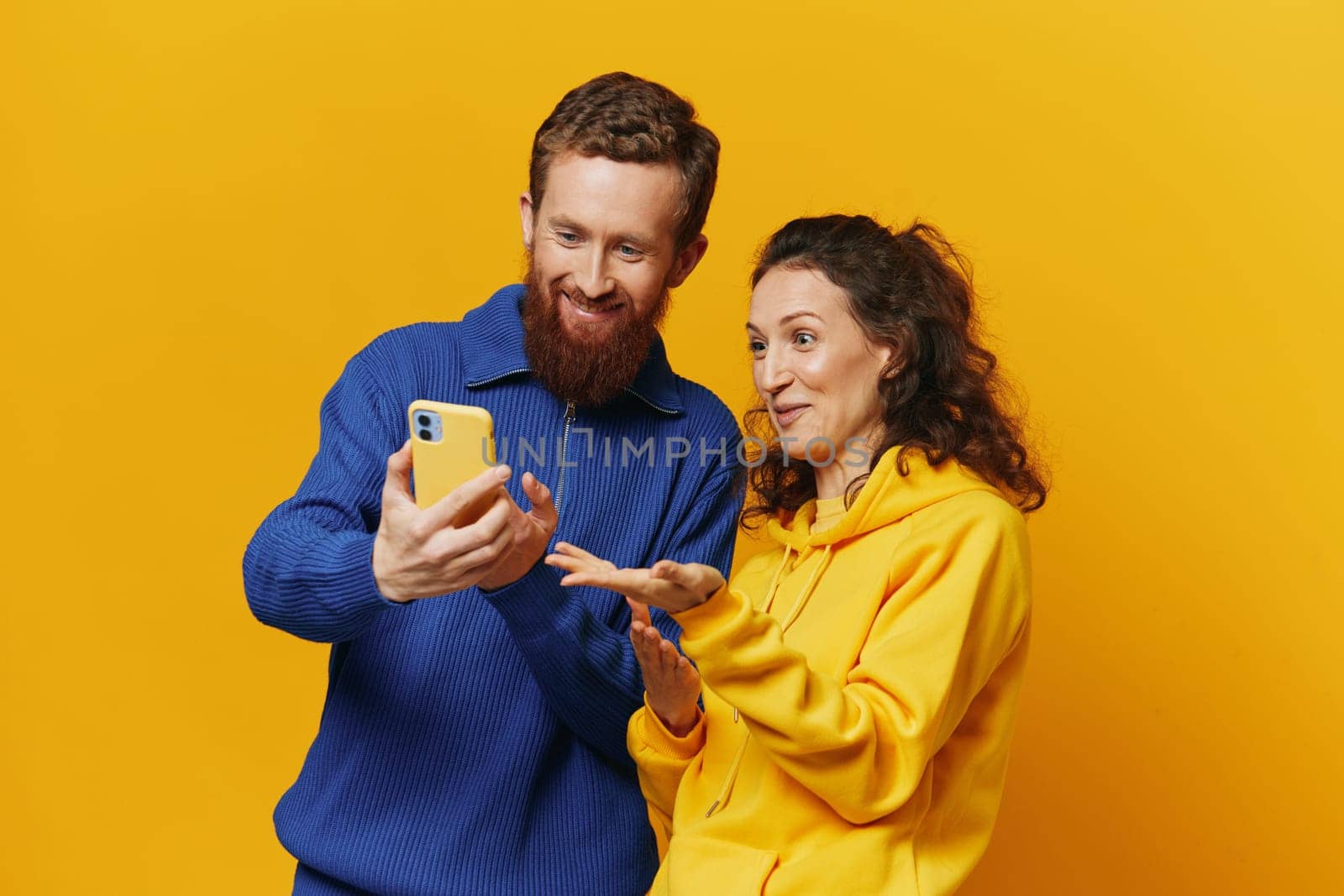 Man and woman couple smiling merrily with phone in hand social media viewing photos and videos, on yellow background, symbols signs and hand gestures, family freelancers. High quality photo