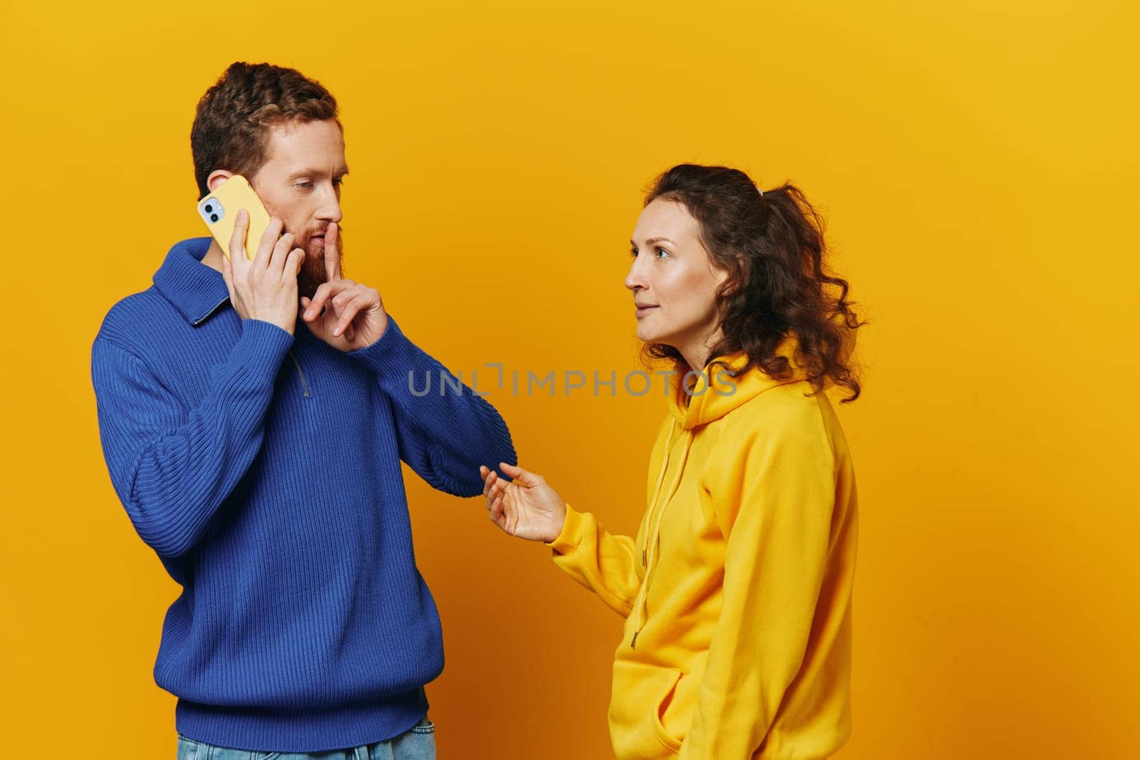 Man and woman couple with phone in hand call talking on the phone, on a yellow background, symbols signs and hand gestures, family quarrel jealousy and scandal. by SHOTPRIME