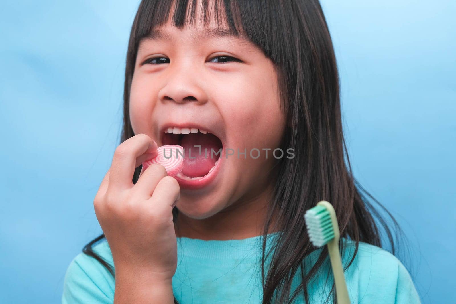 Smiling cute little girl holding toothbrush and sweets isolated on blue background. Kid training oral hygiene and Unhealth eat. Tooth decay prevention or dental care concept. by TEERASAK