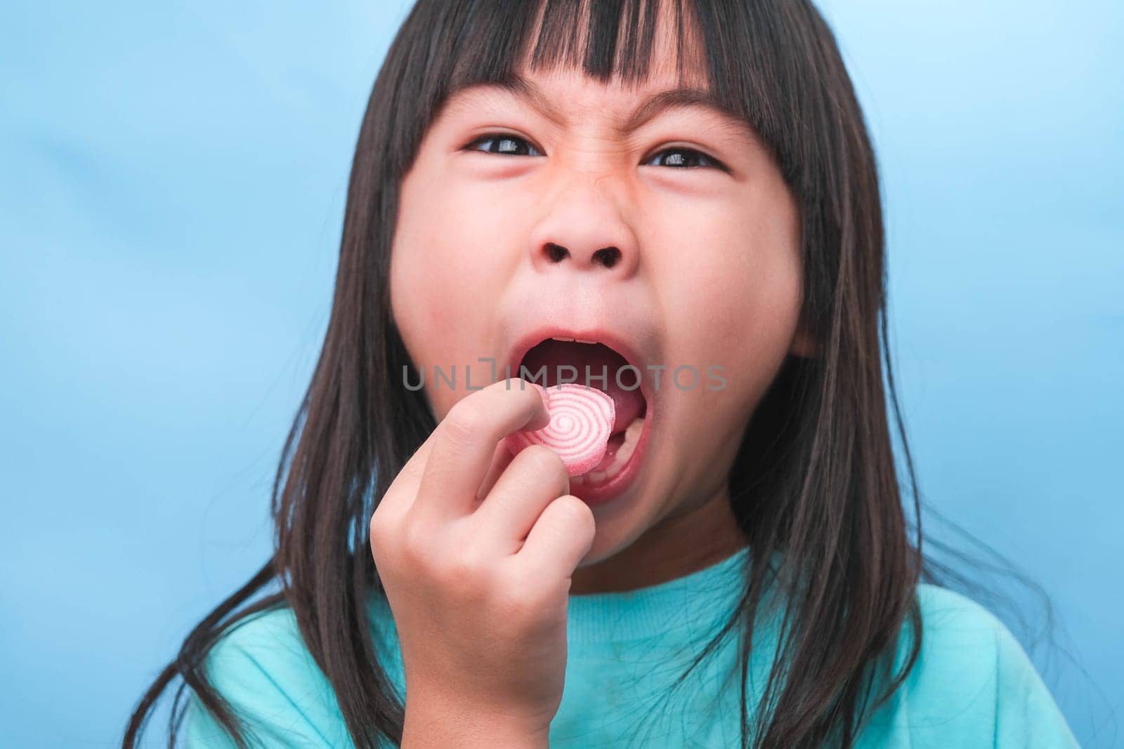 Smiling cute little girl eating sweet gelatin with sugar added isolated on blue background. Children eat sugary sweets, causing loss teeth or tooth decay and unhealthy oral care. by TEERASAK