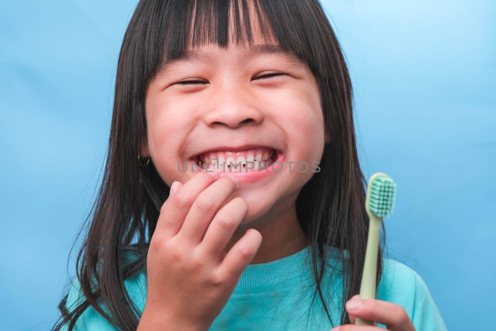 Smiling cute little girl holding toothbrush isolated on blue background. Cute little child brushing teeth. Kid training oral hygiene, Tooth decay prevention or dental care concept. by TEERASAK