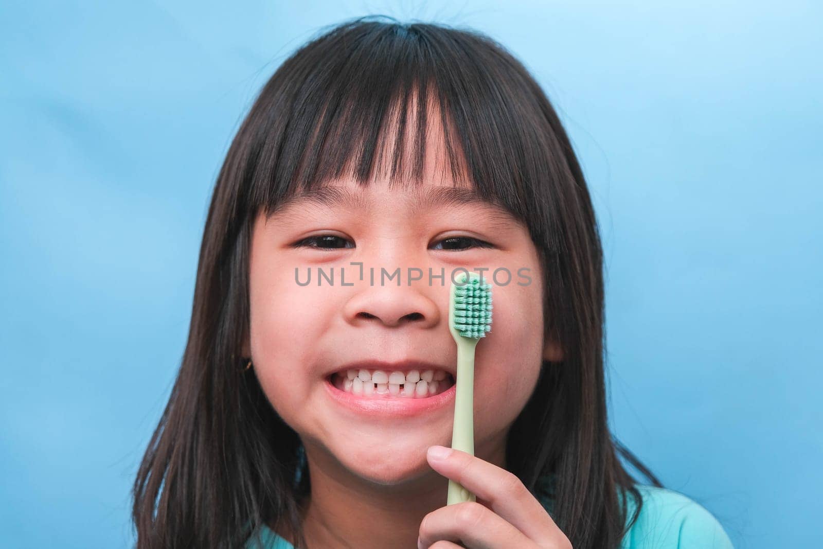 Smiling cute little girl holding toothbrush isolated on blue background. Cute little child brushing teeth. Kid training oral hygiene, Tooth decay prevention or dental care concept. by TEERASAK