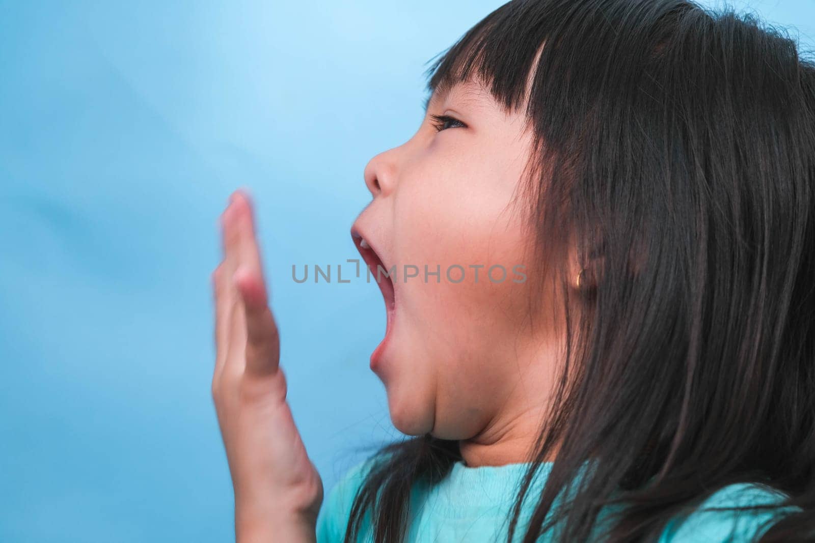 Little asian girl covering her mouth to smell the bad breath. Child girl checking breath with her hands. Oral health problems or dental care concept. by TEERASAK