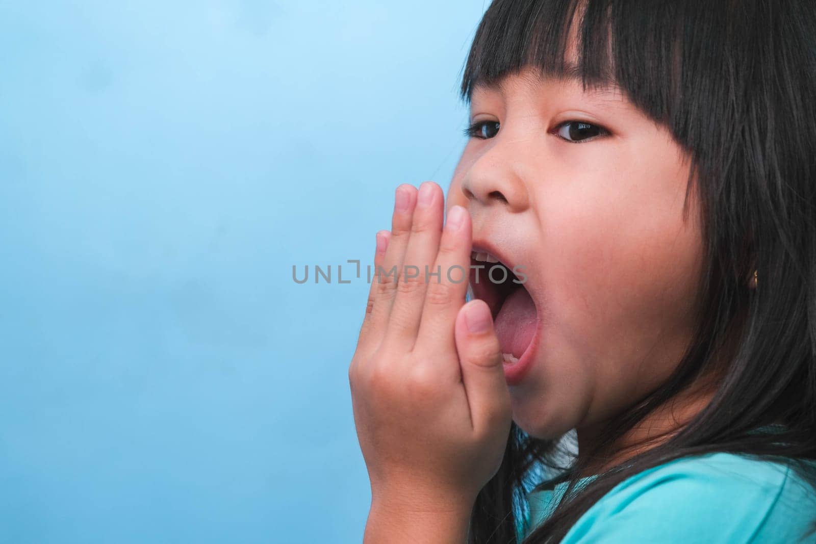 Little asian girl covering her mouth to smell the bad breath. Child girl checking breath with her hands. Oral health problems or dental care concept. by TEERASAK