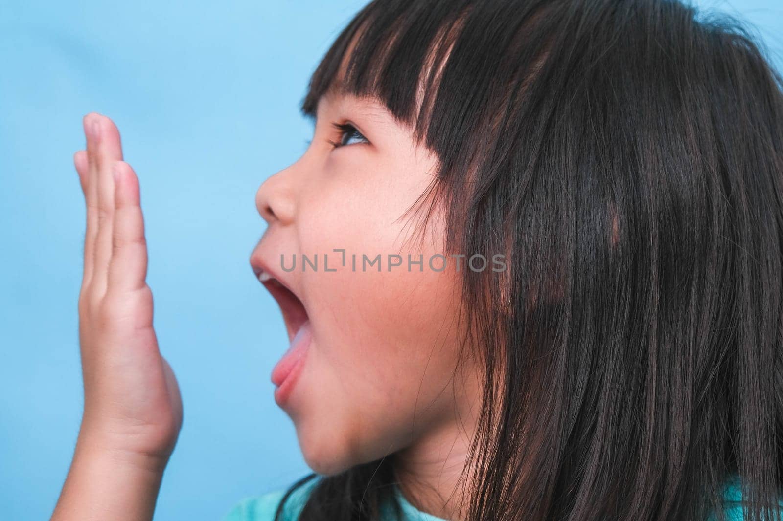 Little asian girl covering her mouth to smell the bad breath. Child girl checking breath with her hands. Oral health problems or dental care concept. by TEERASAK