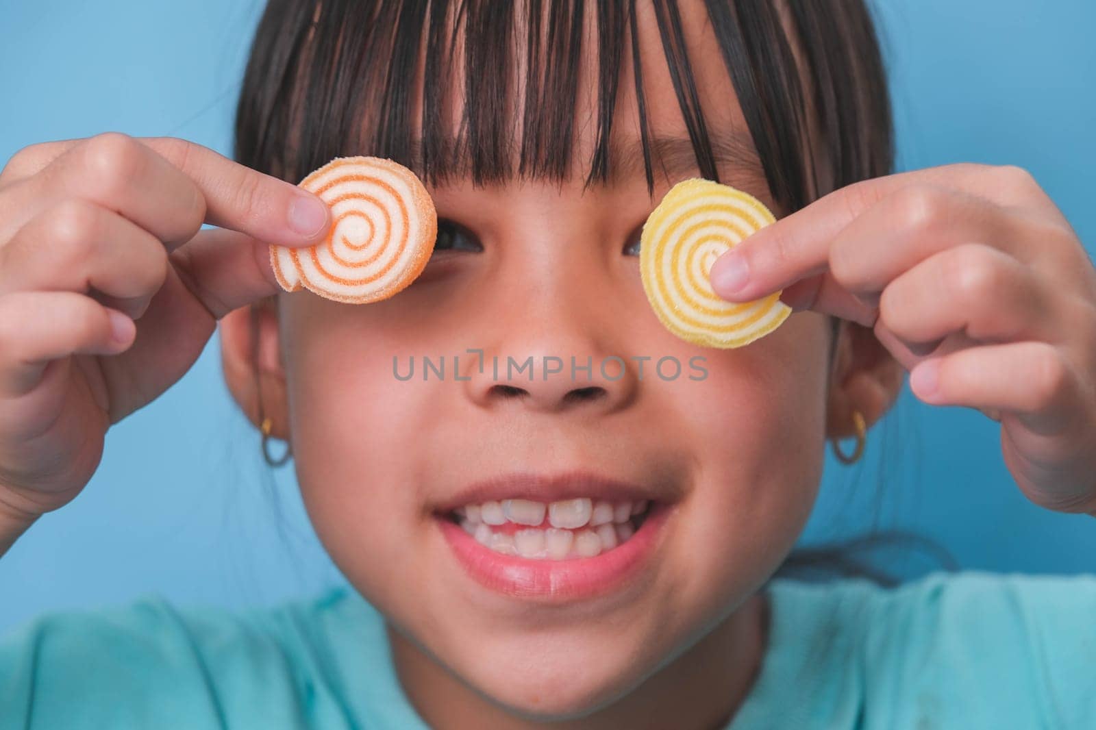Smiling cute little girl covering her eyes with two gelatinous sweets isolated on blue background.  Children eat sugary sweets, causing loss teeth or tooth decay and unhealthy oral care. by TEERASAK