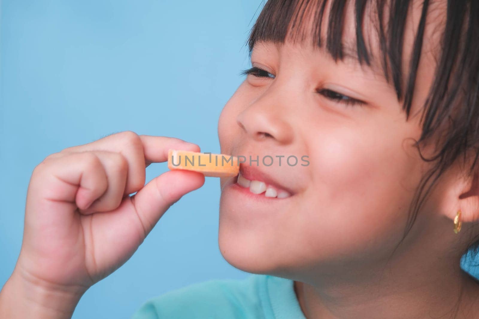 Smiling cute little girl eating sweet gelatin with sugar added isolated on blue background. Children eat sugary sweets, causing loss teeth or tooth decay and unhealthy oral care. by TEERASAK
