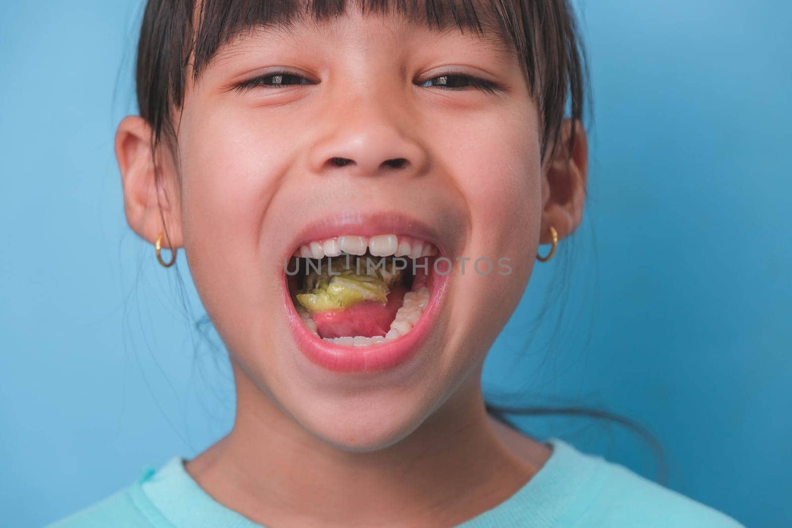 Smiling cute little girl eating sweet gelatin with sugar added isolated on blue background. Children eat sugary sweets, causing loss teeth or tooth decay and unhealthy oral care. by TEERASAK