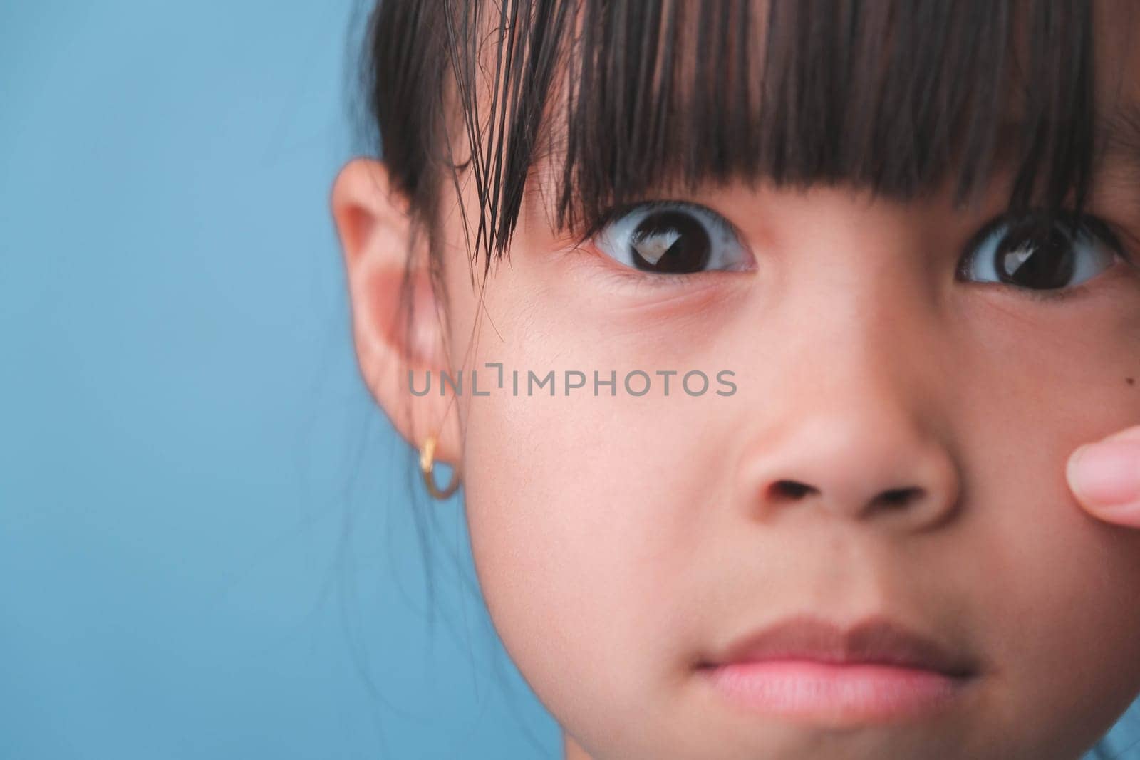 Close-up portrait of surprised young Asian girl isolated on blue background. Cute shocked young girl with big black eyes reacts surprisingly to something, looking at the camera. negative mood child by TEERASAK