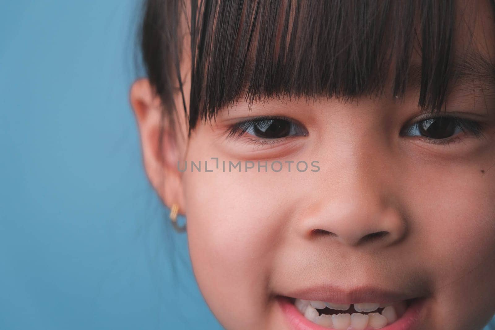 Portrait of a smiling little girl looking at the camera. Close up of cute Asian girl face posing on blue background. by TEERASAK