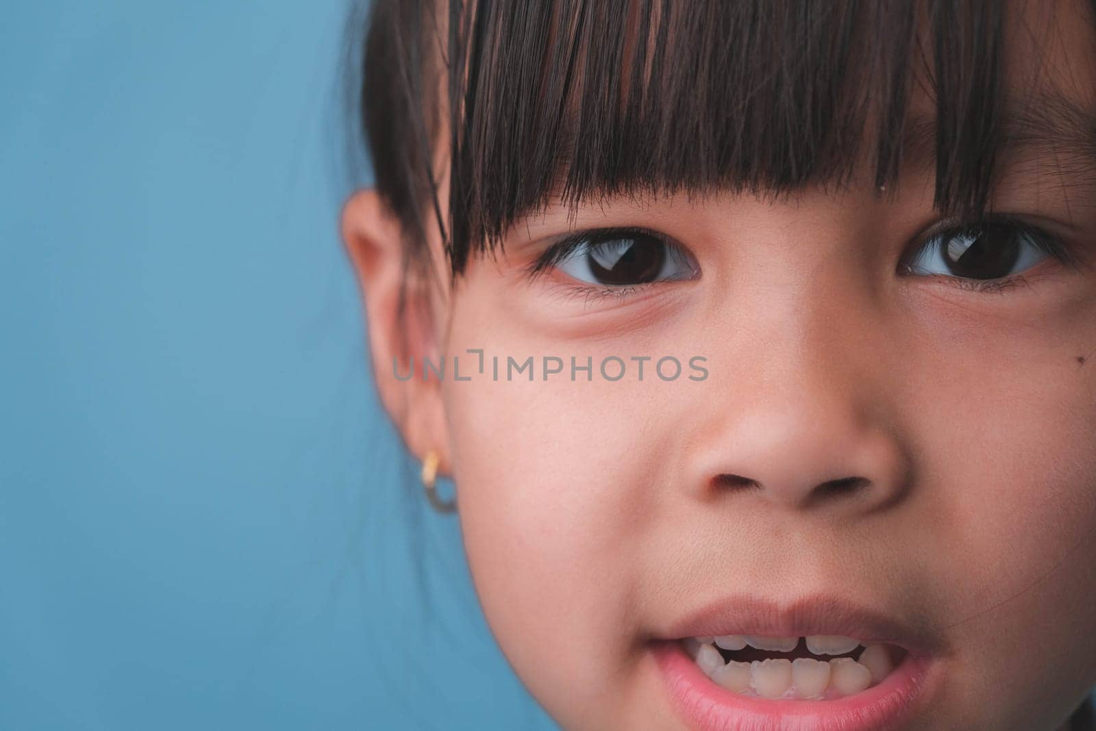Portrait of a smiling little girl looking at the camera. Close up of cute Asian girl face posing on blue background. by TEERASAK