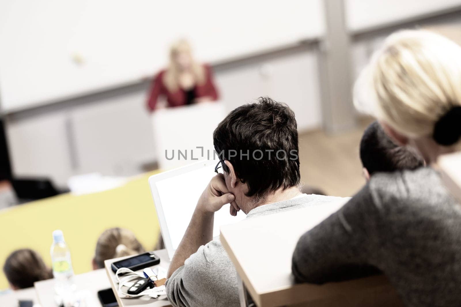 Speaker giving presentation in lecture hall at university. Participants listening to lecture and making notes.