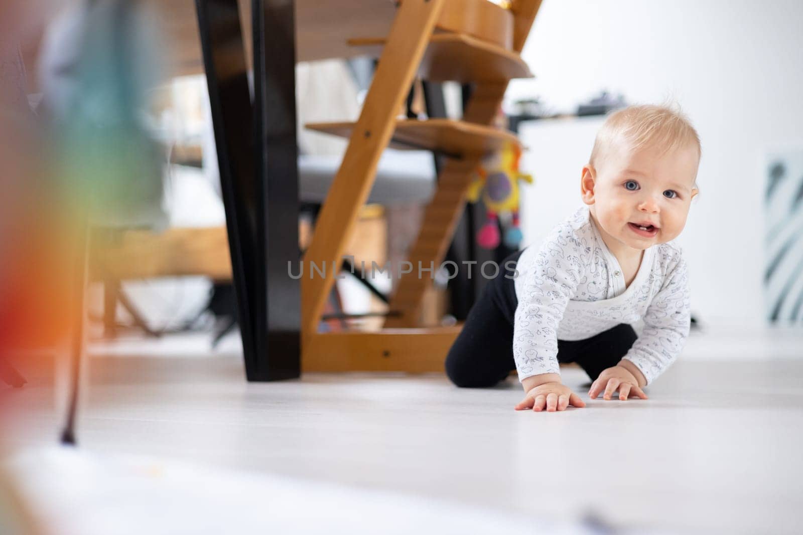 Cute infant baby boy crawling under dining room table at home. Baby playing at home.