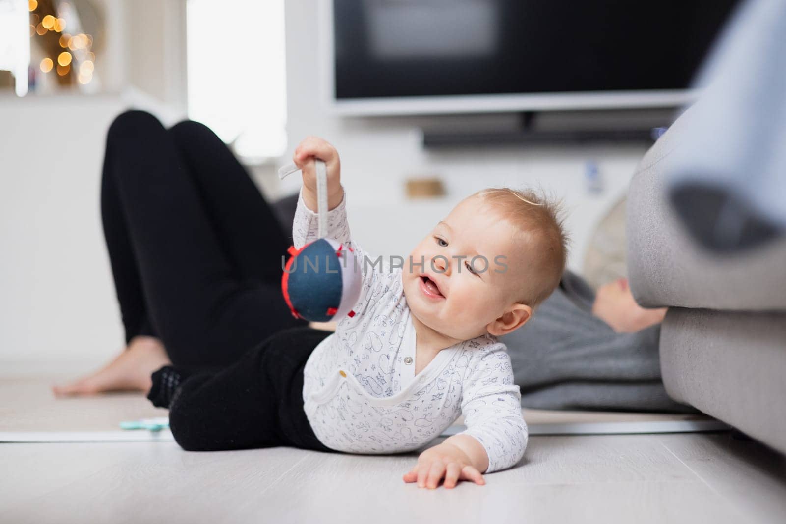 Happy family moments. Mother lying comfortably on children's mat watching and suppervising her baby boy playinghis in living room. Positive human emotions, feelings, joy by kasto