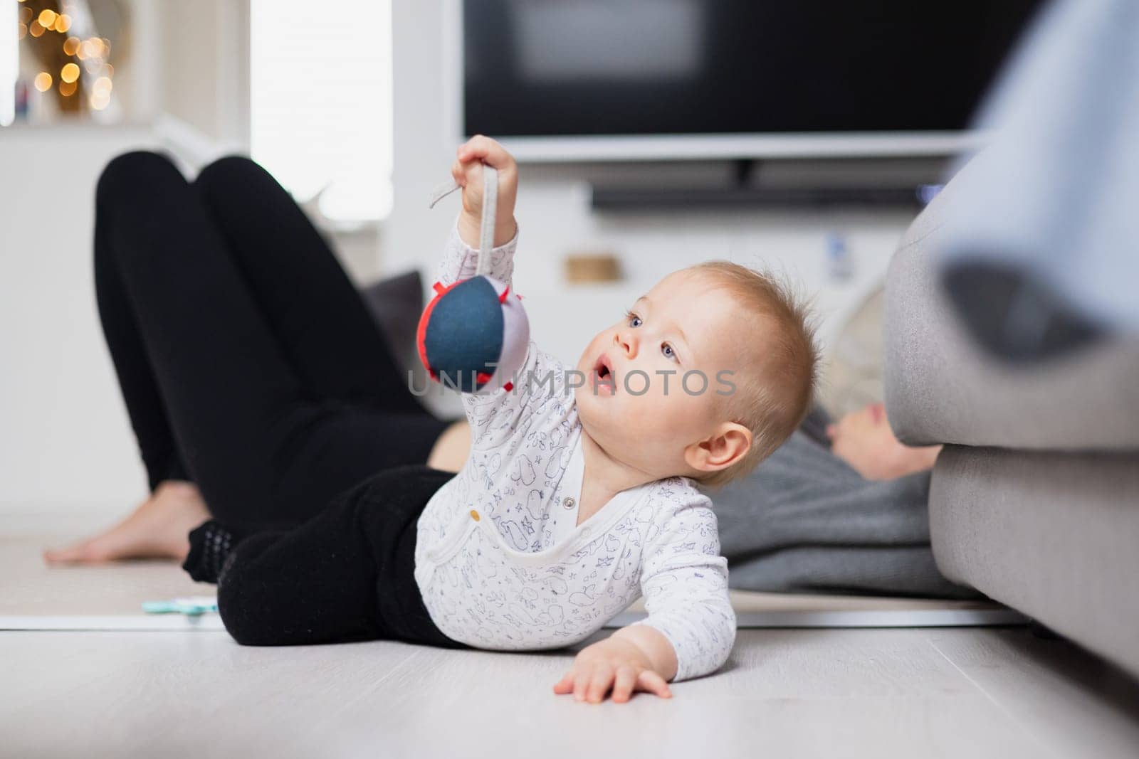 Happy family moments. Mother lying comfortably on children's mat watching and suppervising her baby boy playinghis in living room. Positive human emotions, feelings, joy by kasto