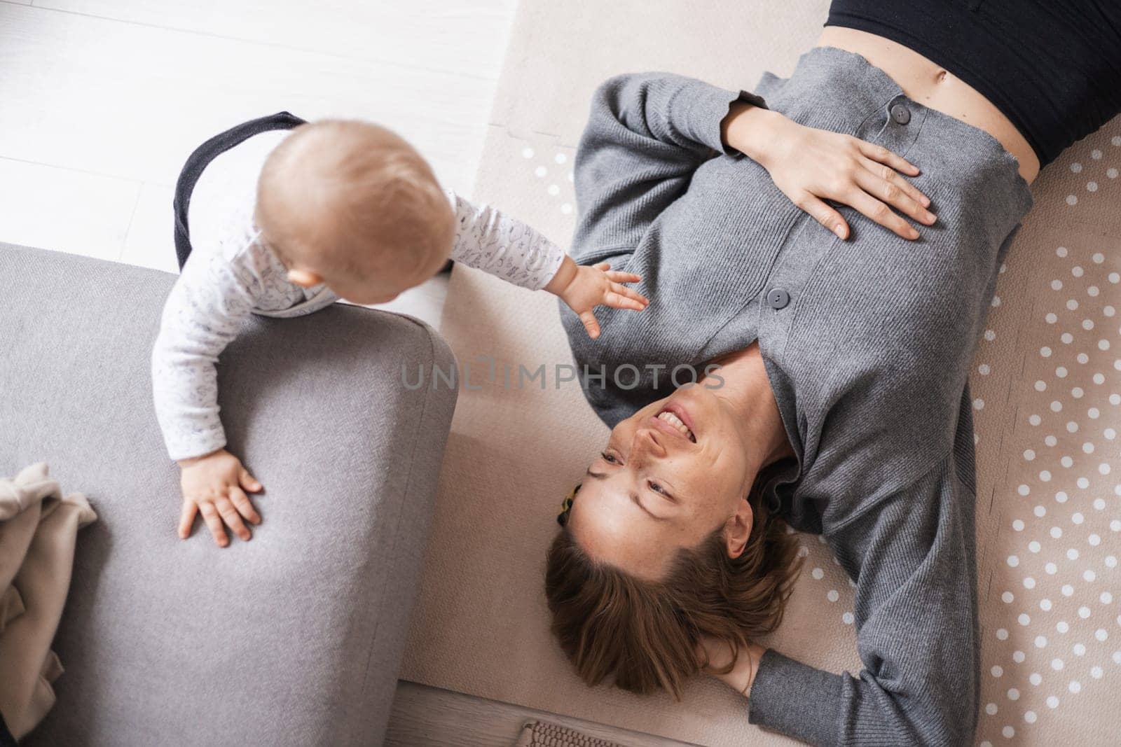 Happy family moments. Mother lying comfortably on children's mat playing with her baby boy watching and suppervising his first steps. Positive human emotions, feelings, joy. by kasto