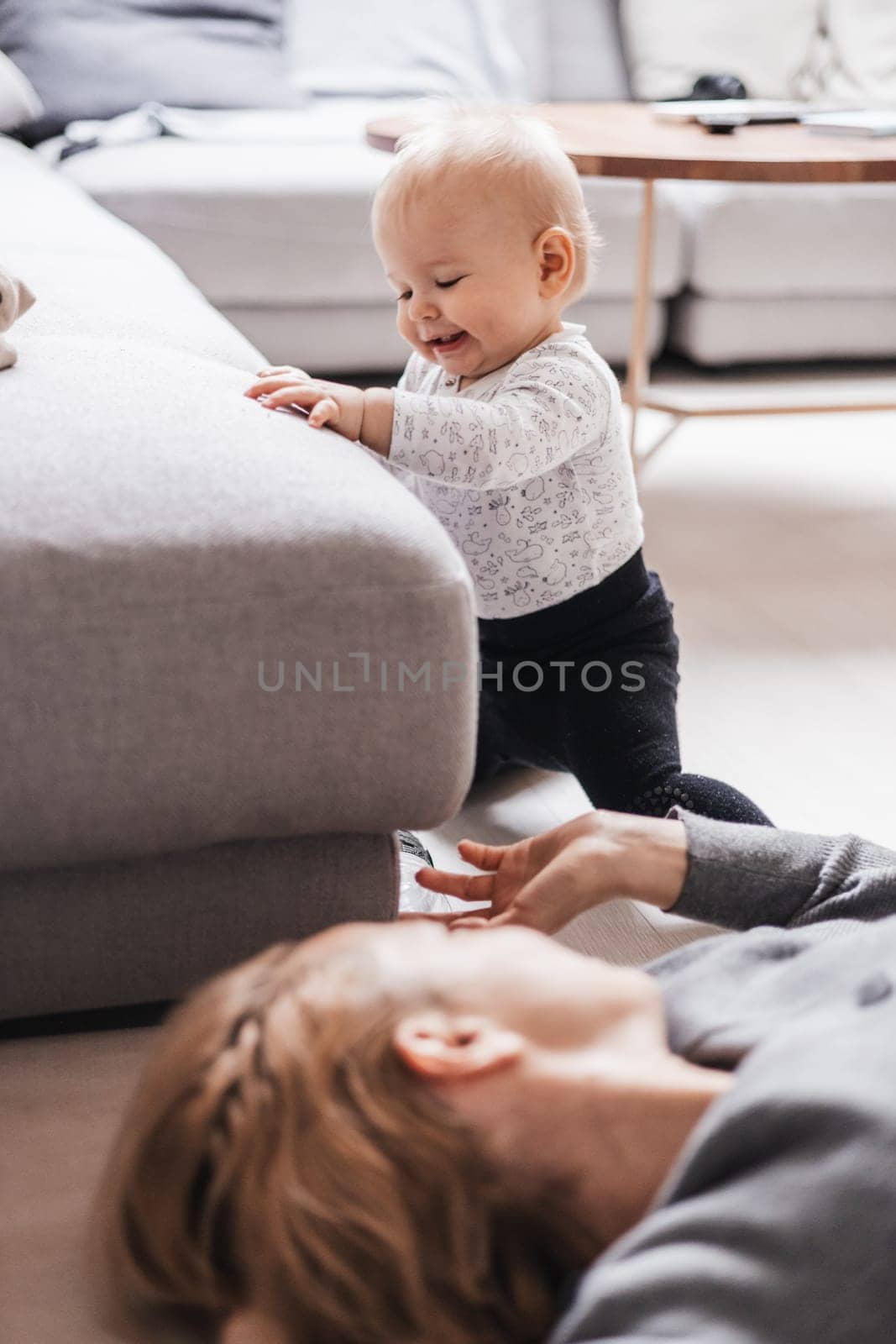 Happy family moments. Mother lying comfortably on children's mat playing with her baby boy watching and suppervising his first steps. Positive human emotions, feelings, joy