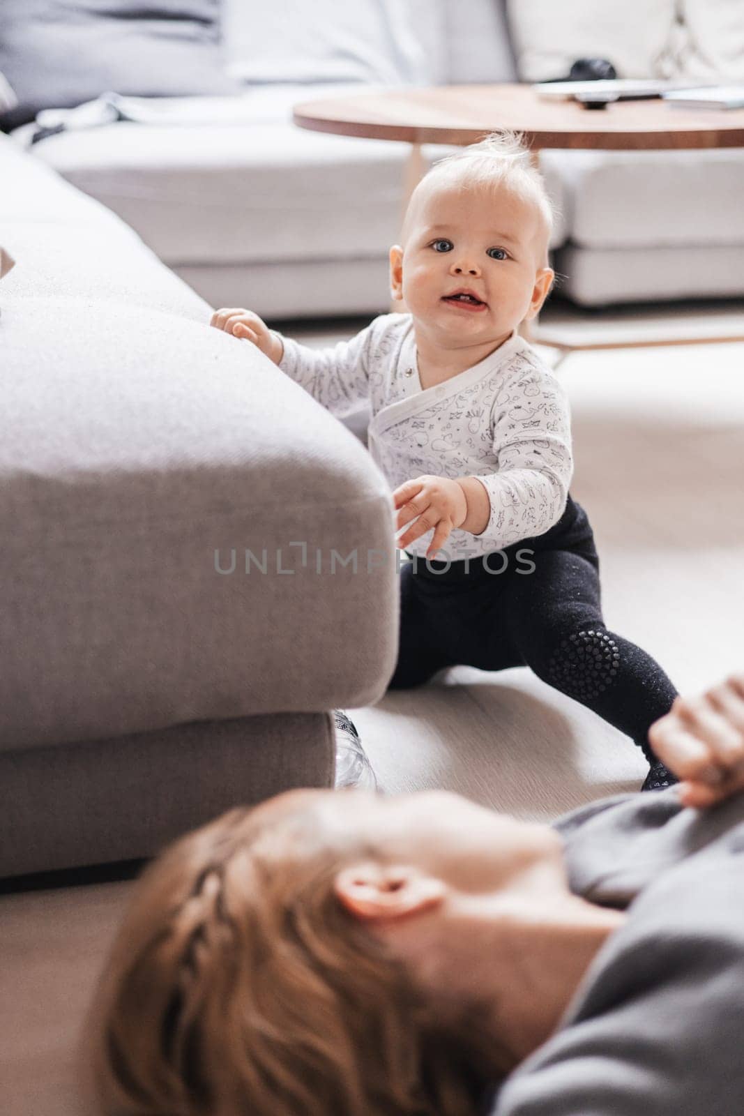Happy family moments. Mother lying comfortably on children's mat playing with her baby boy watching and suppervising his first steps. Positive human emotions, feelings, joy