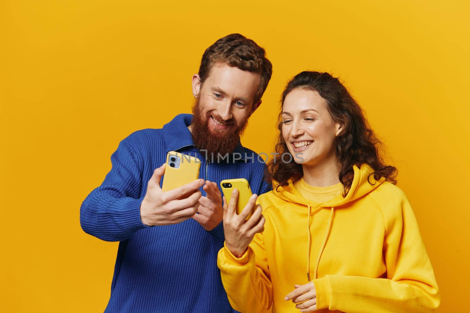 Man and woman couple smiling merrily with phone in hand social media viewing photos and videos, on yellow background, symbols signs and hand gestures, family freelancers. High quality photo