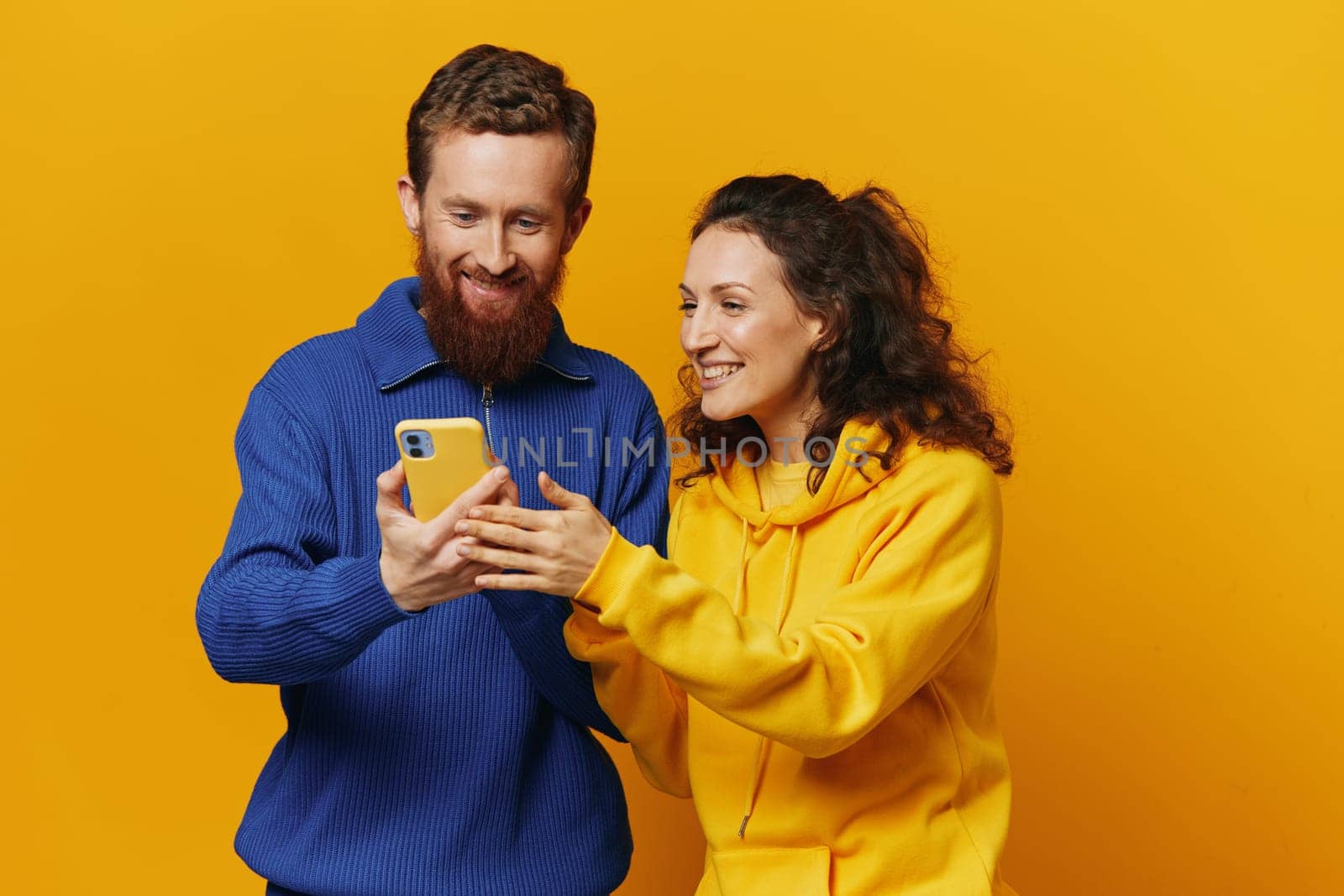 Man and woman couple smiling merrily with phone in hand social media viewing photos and videos, on yellow background, symbols signs and hand gestures, family freelancers. High quality photo