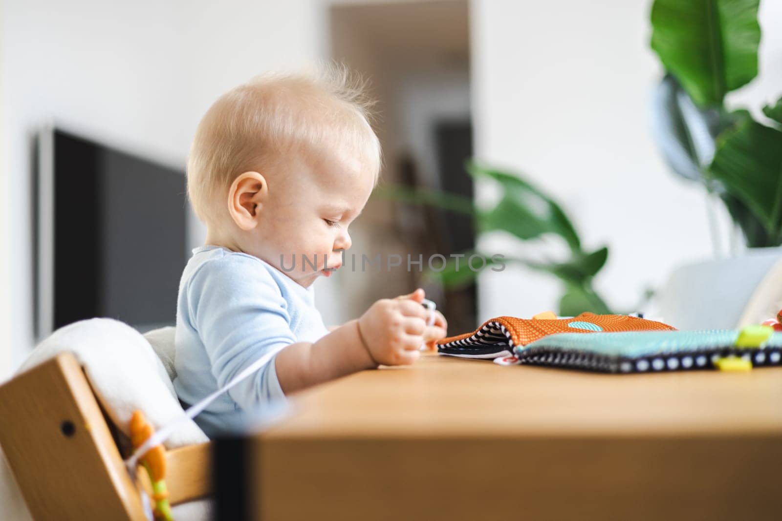 Happy infant sitting at dining table and playing with his toy in traditional scandinavian designer wooden high chair in modern bright atic home. Cute baby playing with toys.