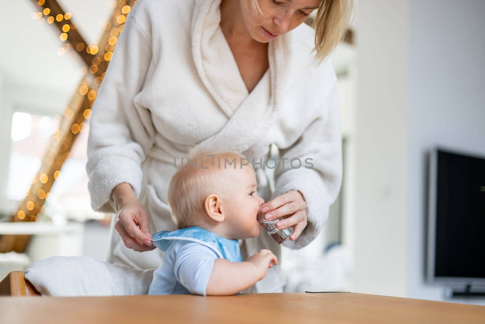 Casual domestic lifestyle moments concept. Infant baby boy drinking water from small glass with a help of his mother wearing cosy bathrope