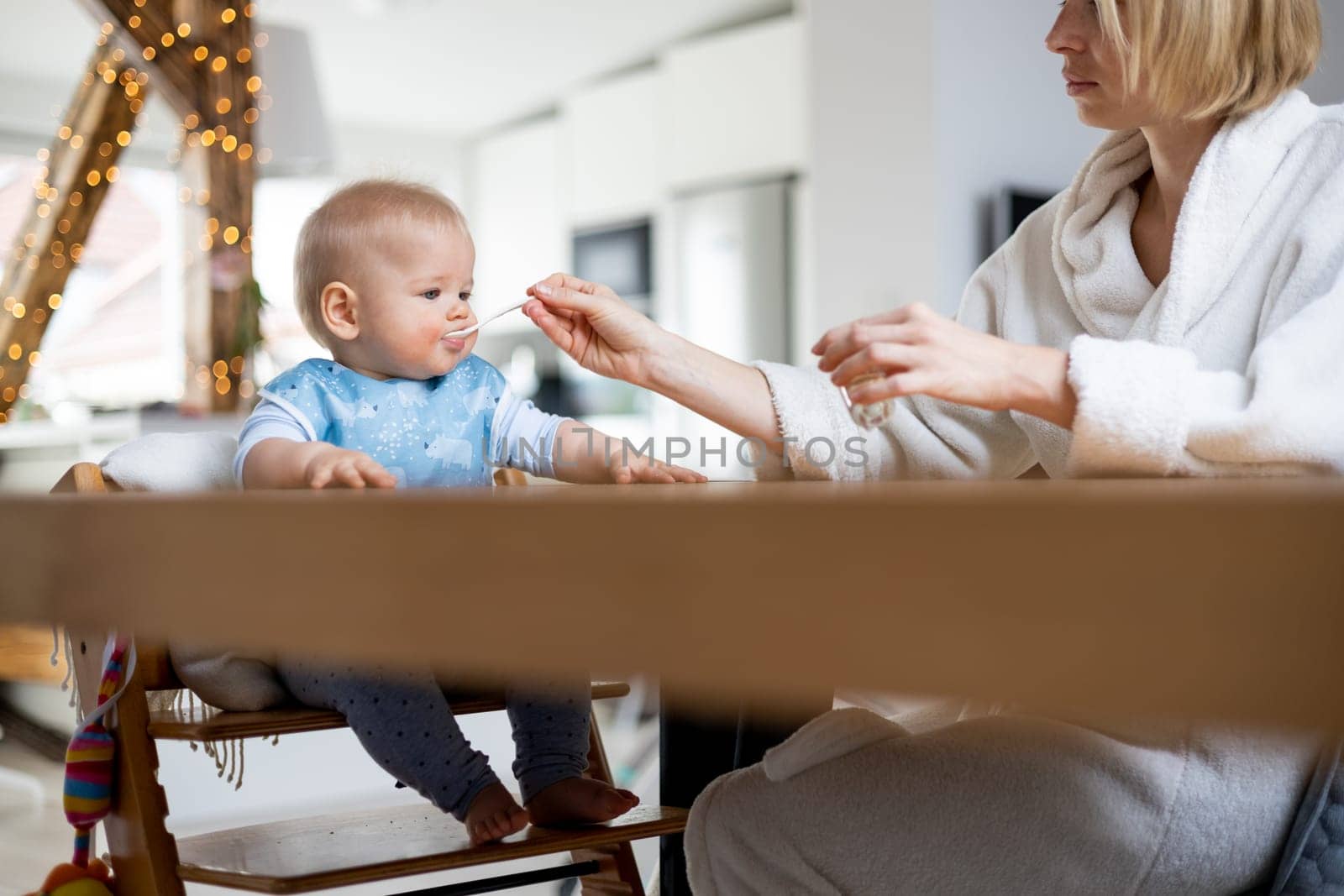 Mother wearing cosy bathrope spoon feeding her baby boy child in baby high chair with fruit puree at dinning table at home. Baby solid food introduction concept.