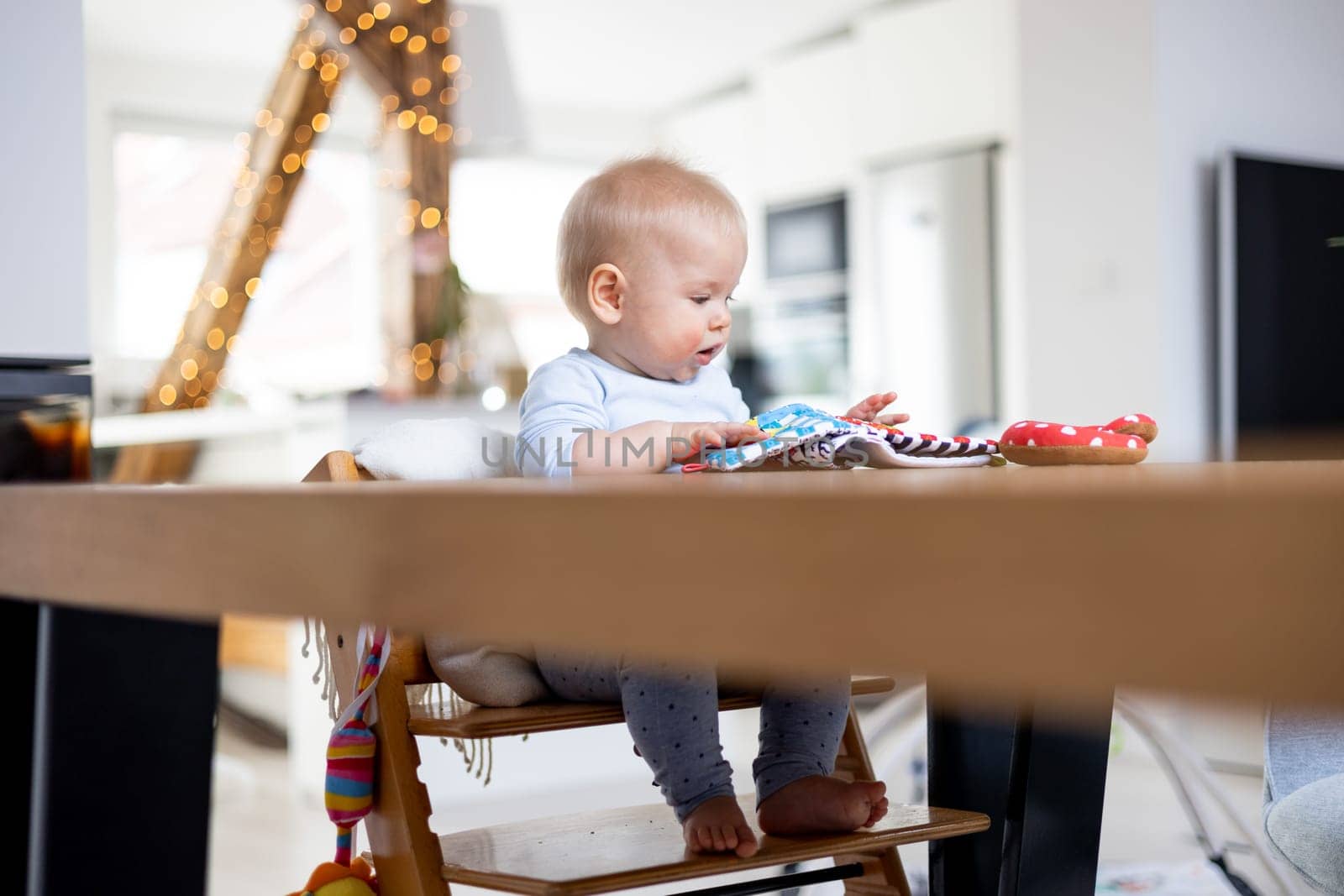 Happy infant sitting at dining table and playing with his toy in traditional scandinavian designer wooden high chair in modern bright atic home. Cute baby playing with toys.