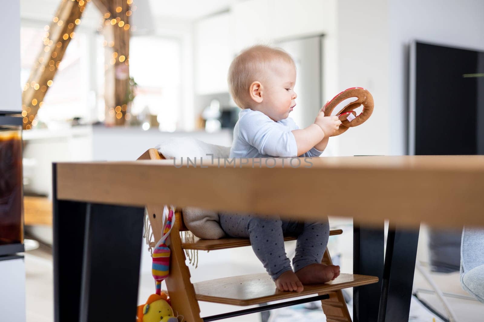 Happy infant sitting at dining table and playing with his toy in traditional scandinavian designer wooden high chair in modern bright atic home. Cute baby playing with toys.