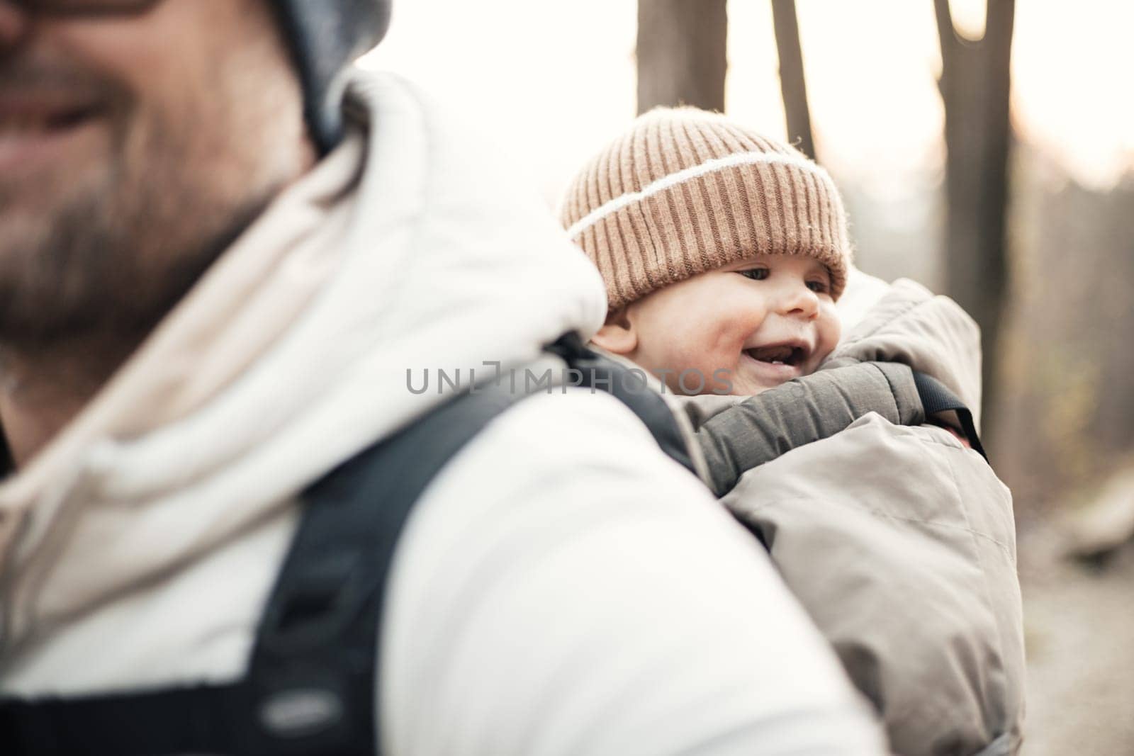 Sporty father carrying his infant son wearing winter jumpsuit and cap in backpack carrier hiking in autumn forest