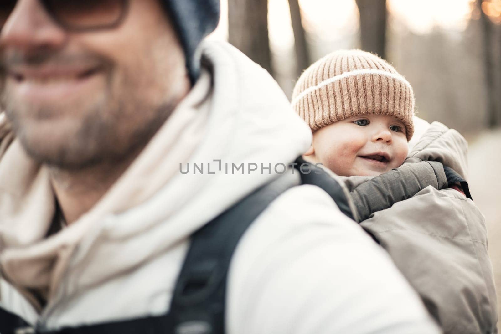 Sporty father carrying his infant son wearing winter jumpsuit and cap in backpack carrier hiking in autumn forest