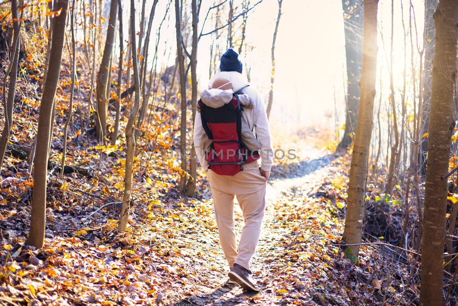 Sporty father carrying his infant son wearing winter jumpsuit and cap in backpack carrier hiking in autumn forest