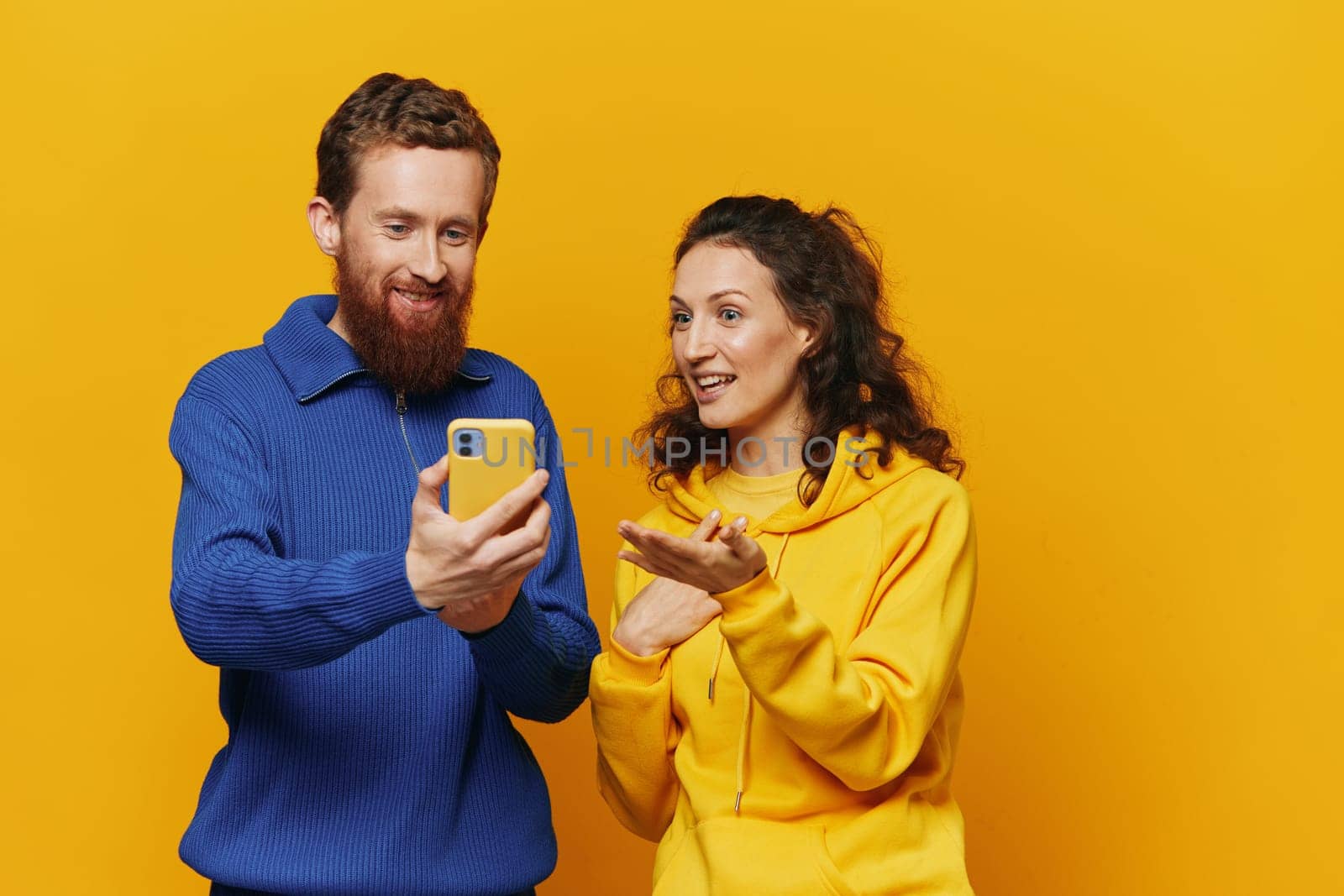 Man and woman couple smiling merrily with phone in hand social media viewing photos and videos, on yellow background, symbols signs and hand gestures, family freelancers. High quality photo