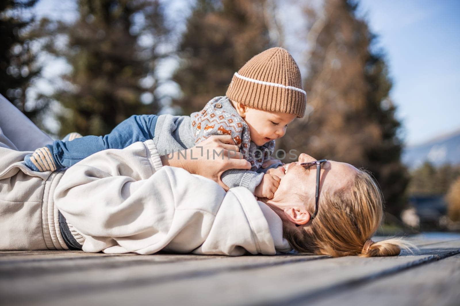 Happy family. Young mother playing with her baby boy infant oudoors on sunny autumn day. Portrait of mom and little son on wooden platform by lake. Positive human emotions, feelings, joy