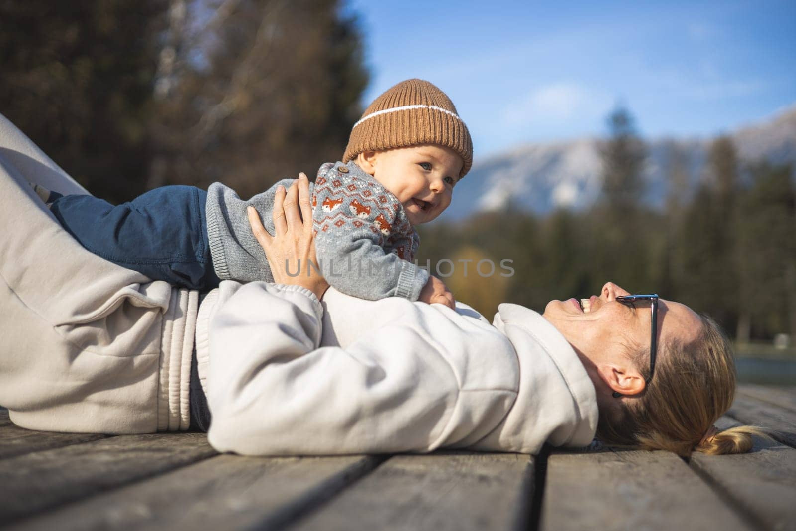 Happy family. Young mother playing with her baby boy infant oudoors on sunny autumn day. Portrait of mom and little son on wooden platform by lake. Positive human emotions, feelings, joy
