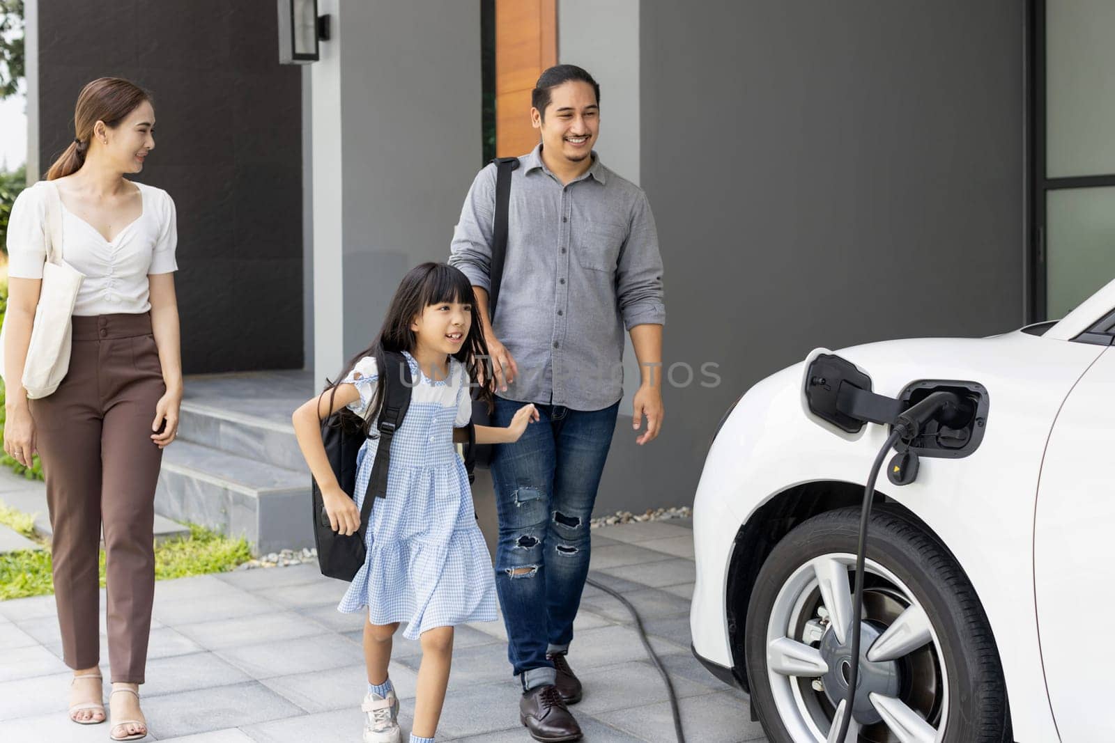 Progressive young parents and daughter with electric vehicle and home charging station. Green and clean energy from electric vehicles for healthy environment. Eco power from renewable source at home.