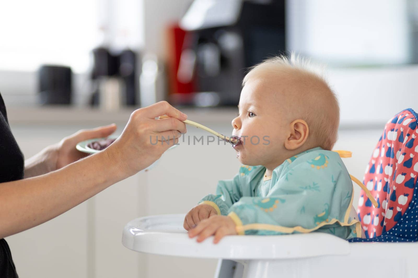 Mother spoon feeding her baby boy child in baby chair with fruit puree in kitchen at home. Baby solid food introduction concept