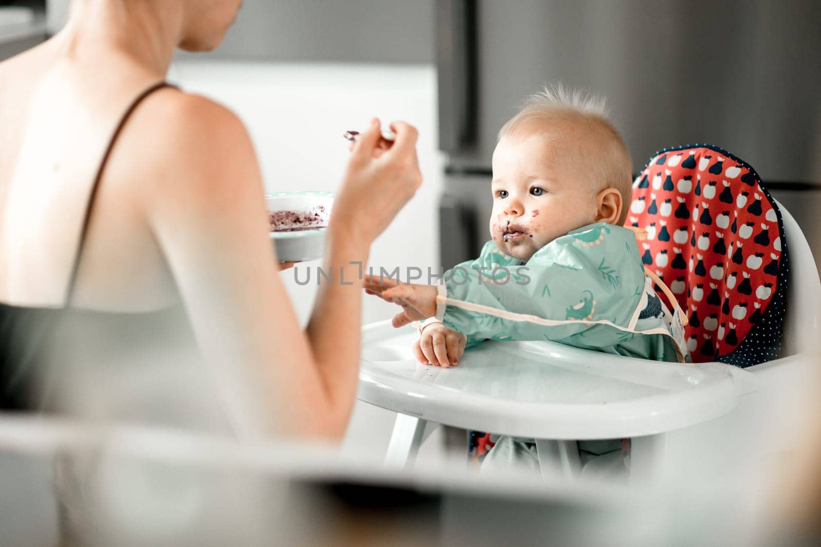 Mother spoon feeding her baby boy child in baby chair with fruit puree in kitchen at home. Baby solid food introduction concept. by kasto