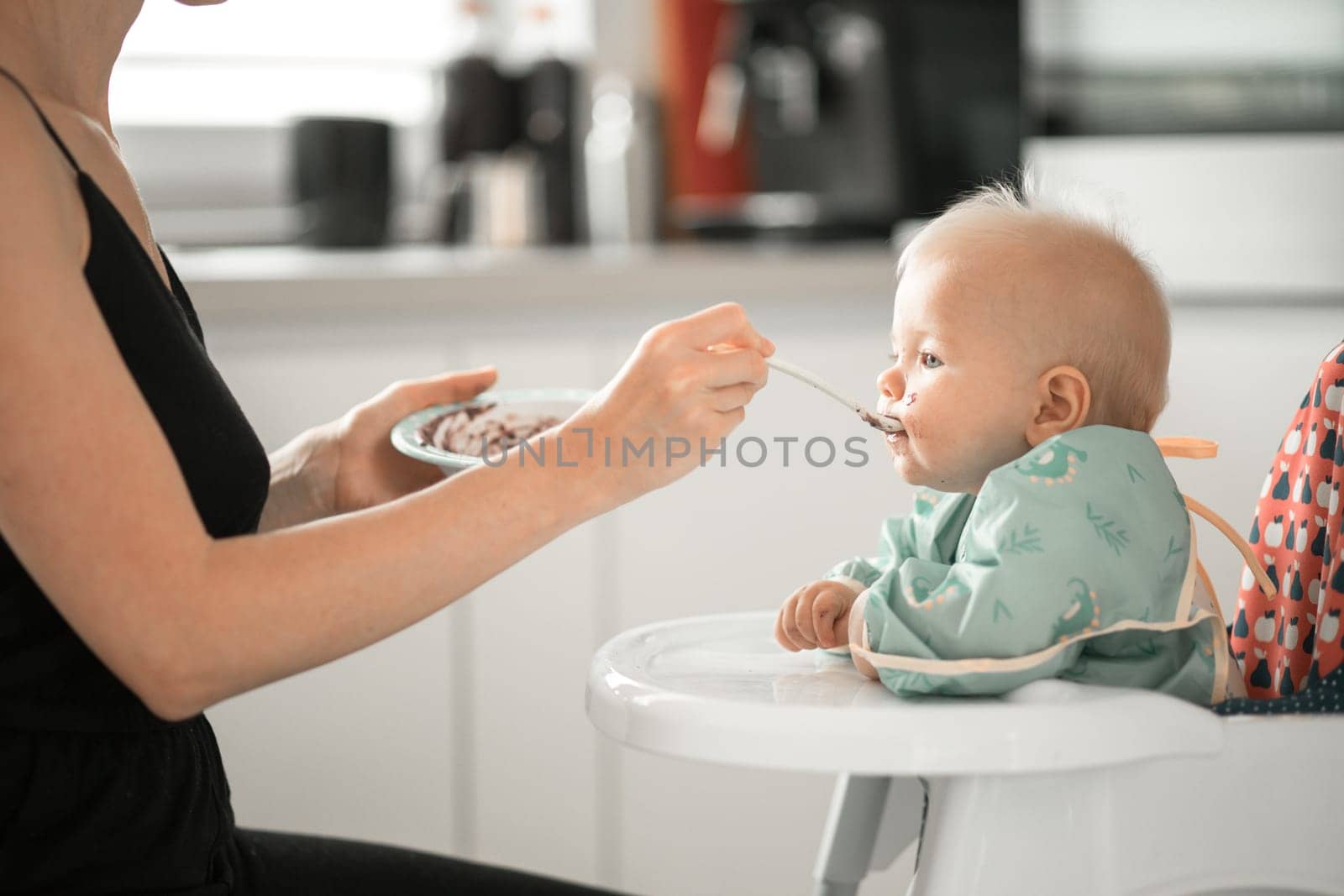 Mother spoon feeding her baby boy child in baby chair with fruit puree in kitchen at home. Baby solid food introduction concept